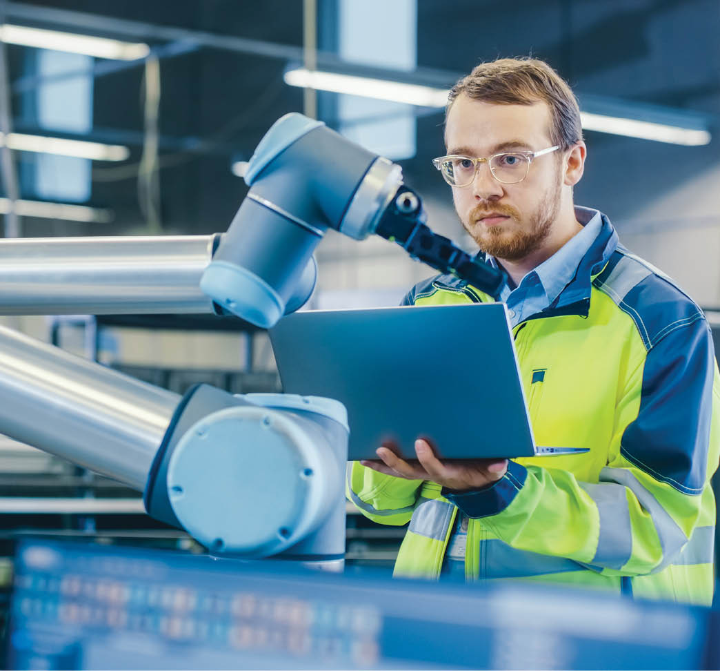 At the Factory: Automation Engineer Uses Laptop for Programming Robotic Arm. New Era in Automatic Manufacturing Industry.