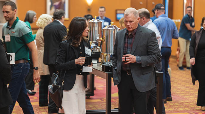 A group of people are gathered in a room, some of them are holding drinks. A man and a woman are standing near a coffee urn, engaged in conversation. There are other people in the room, some of them wearing ties. The room appears to be a convention or a social event. AI generated content
