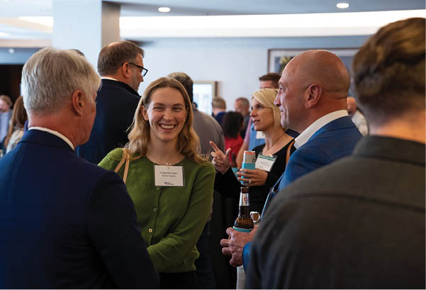 A group of people are standing in a room, socializing and enjoying beverages. A woman is smiling and holding a beer bottle. AI generated content