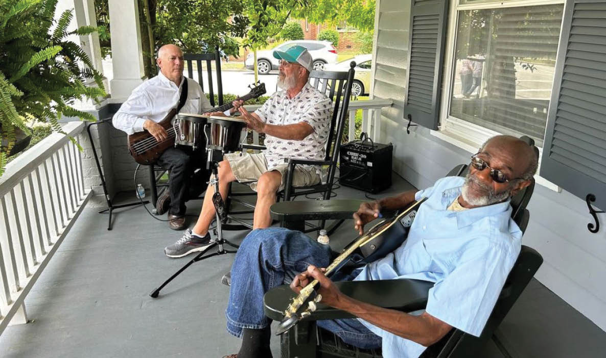 Three men playing guitars on a porch. Description generated by AI