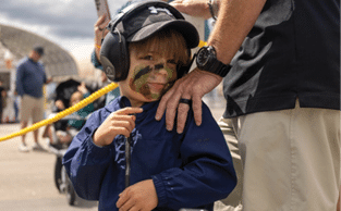 A young boy wearing a hat and camouflage paint on his face. Description generated by AI