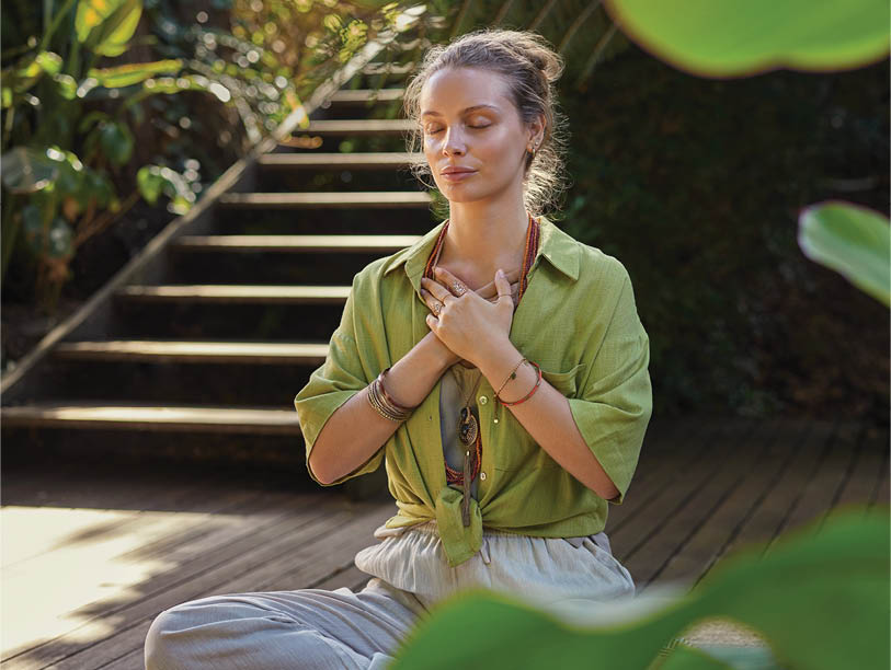 Peaceful woman sits cross-legged in a meditative state, hands over her heart, expressing gratitude and mindfulness in a lush. Girl connects with her emotions during a reflective moment, embracing inner peace. Calm woman practices a heart-centered meditation, cultivating self-love and healing energy, surrounded by greenery.