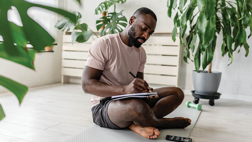 Young African-American man sitting cross-legged on a yoga mat at home, writing in a notebook after a workout, with a smartphone nearby, creating a calm and focused atmosphere.