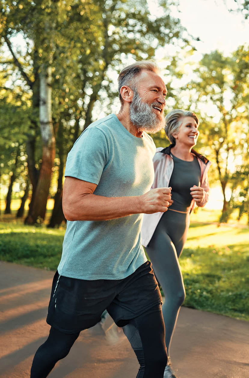 Cardio training outdoors. Side view of caucasian grey bearded man and beautiful old woman running along summer park with sunlight on background. Cheerful retired people leading active lifestyle.
