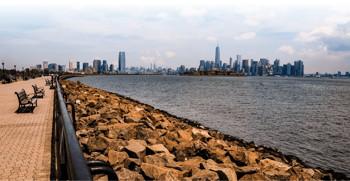 A view of the skyline of Jersey City to the Left and Lower Manhattan on the right from Liberty State Park in New Jersey on a bright sunny day with the waters of the Hudson River in the foreground and the walkway of the park to the left. Laying in front of Lower Manhattan is Ellis Island, famous for being the entry point of thousands of immigrants that arrived to the United States in the early Twentieth Century.