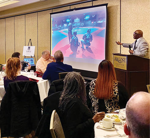 A man is giving a presentation in front of a group of people. The audience is seated at tables with plates, cups, and utensils. The man is standing at a podium, and a large screen is visible behind him. AI generated content
