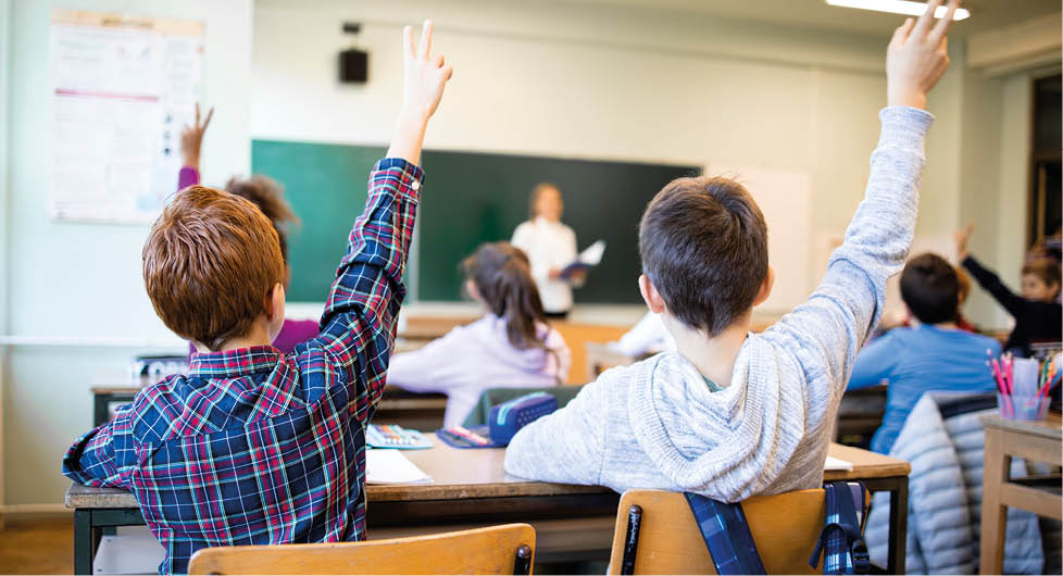 Schoolchildren at classroom with raised hands answering teacher's question.