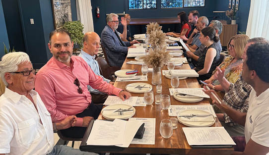A group of people are sitting around a wooden table with plates of food and drinks. Description generated by AI