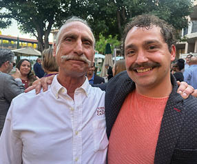 Two men standing together, one with a mustache and the other with a beard. Description generated by AI