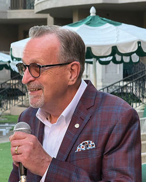 A man wearing a suit and tie is speaking into a microphone. Description generated by AI