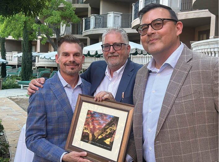Three men are standing together, two of them wearing suits and ties. Description generated by AI