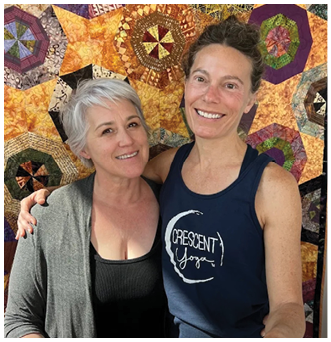 Two women standing in front of a quilt. Description generated by AI