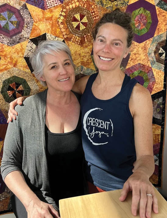 Two women standing in front of a quilt. Description generated by AI