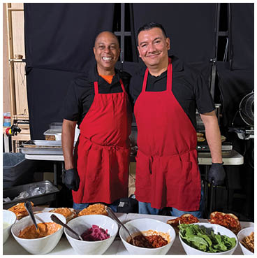Two men wearing red aprons and smiling. Description generated by AI