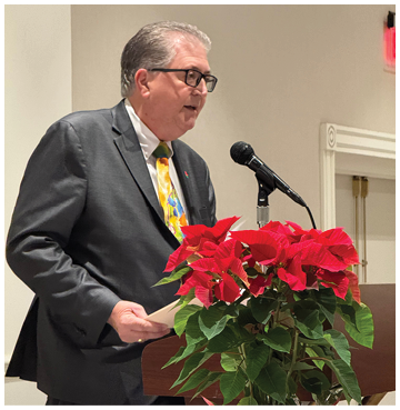A man in a gray suit is speaking at a podium with a red flower in a vase in front of him. Description generated by AI