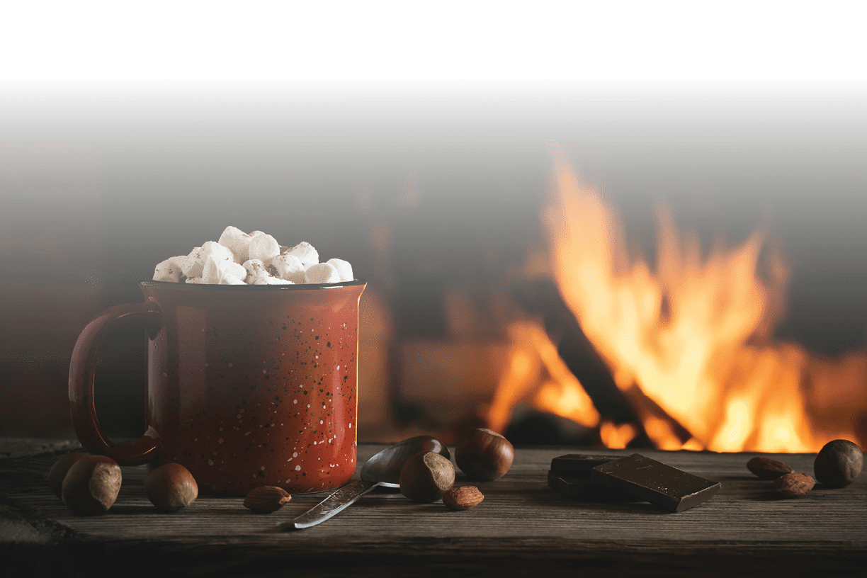 Cocoa with marshmallows and chocolate in a red mug on a wooden table near a burning fireplace.