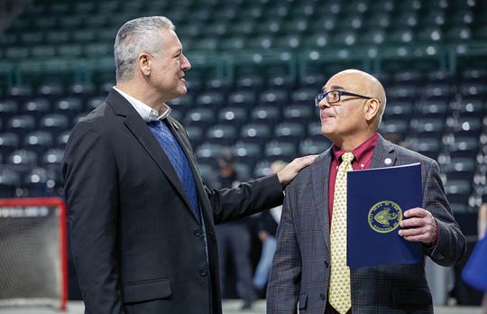 Two men are standing in a stadium, one holding a blue folder. AI generated content