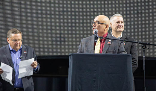 Three men stand on stage, each holding a piece of paper. They are wearing suits and ties, and one of them is wearing a red tie. The men appear to be giving a presentation or speech. AI generated content