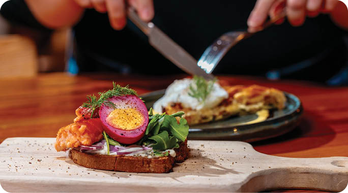 A person is cutting food on a plate with a knife and fork. The plate contains a sandwich, a pickle, and a slice of pizza. The person is sitting at a wooden table. AI generated content