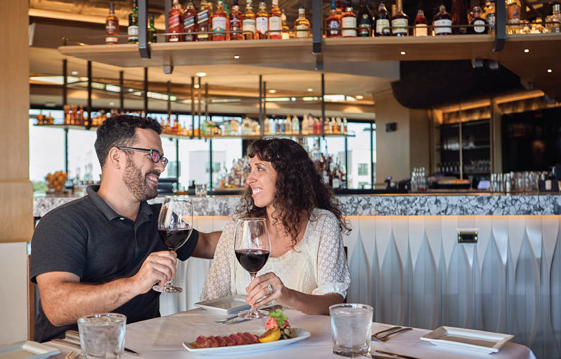 A man and woman are sitting at a table in a restaurant, enjoying a meal together. They are holding wine glasses and a bottle of wine is on the table. The table is set with silverware, including forks, knives, and spoons. There are also cups and bowls on the table. The restaurant has a bar with numerous bottles and wine glasses displayed. AI generated content