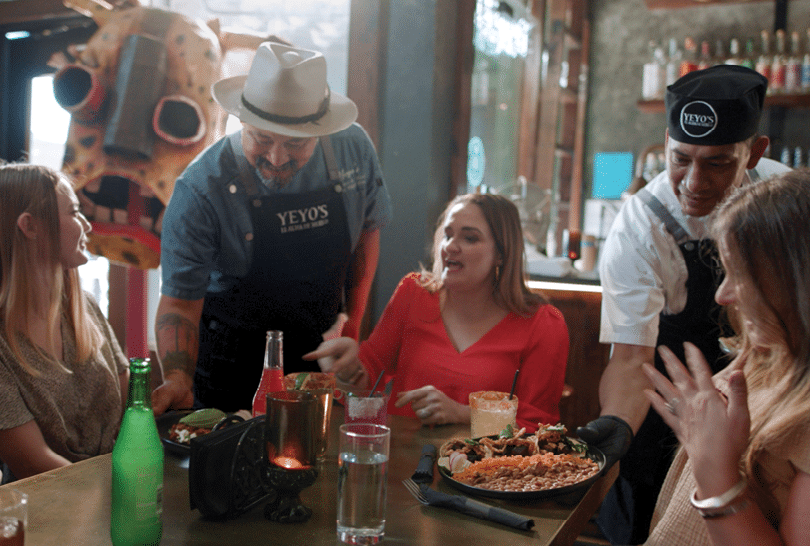 A group of people are sitting around a table with food and drinks. A man in a cowboy hat is serving them. There is a stuffed animal on the table. AI generated content