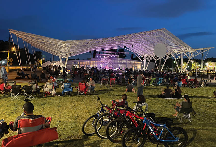A large crowd of people are sitting on the grass in front of a stage, watching a concert. There are several bicycles parked nearby, and a few chairs are scattered throughout the area. The event is taking place at night, and the atmosphere is lively. AI generated content