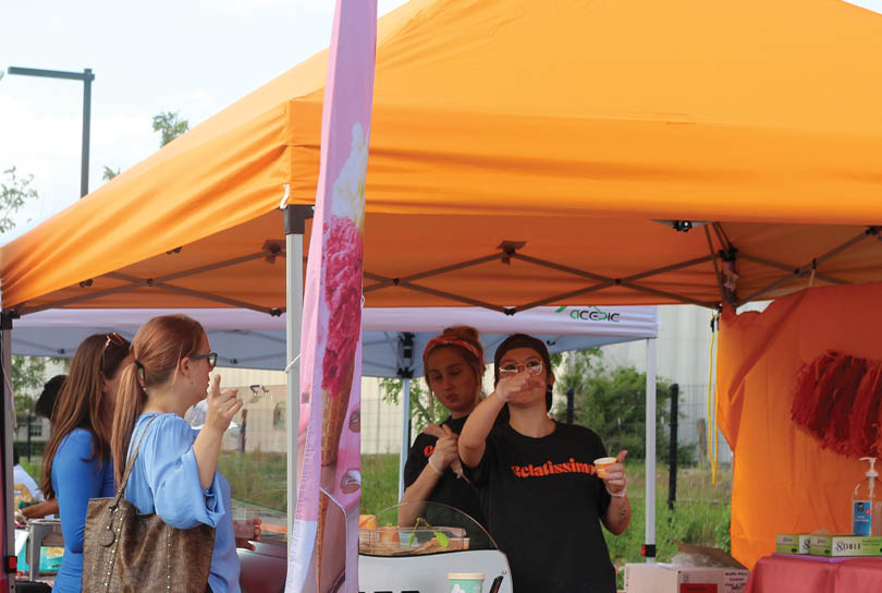 A group of people are gathered around a table with food and drinks. They are standing in line to get their food and drinks. There are several people in the line, and they are all waiting for their turn to get their food and drinks. AI generated content