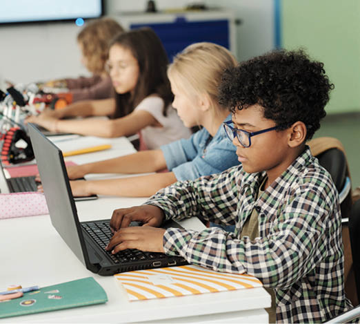 Side view of youthful African American schoolboy and his classmates working in front of laptops while sitting in row by desk