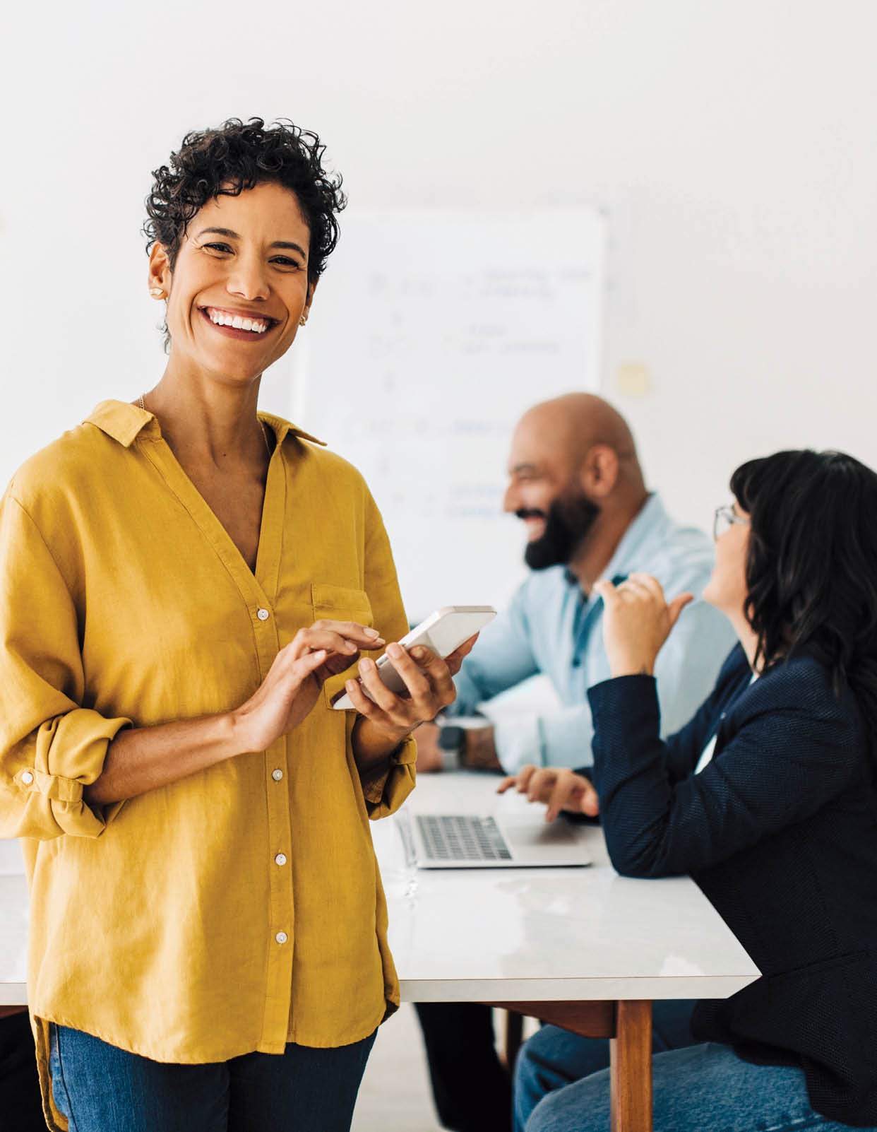 Business woman standing in a boardroom and using a smart phone. Black female professional smiling at the camera in an office. Colleagues having a meeting in the background.