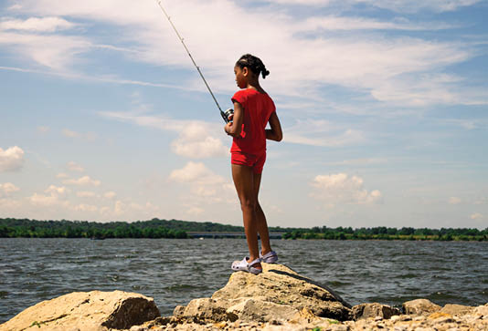 A woman in a red shirt is standing on a rock near a body of water, holding a fishing rod. AI generated content