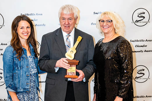 Three people are standing in front of a wall, with one man holding a trophy. The woman on the left is wearing a blue shirt and a floral skirt. The woman on the right is wearing a black dress and glasses. The man in the middle is holding a trophy and wearing a suit. AI generated content