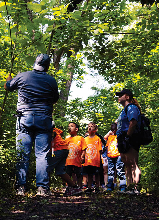 A group of people, including both men and women, are standing in a wooded area. They are wearing orange shirts and appear to be listening to a man who is speaking to them. Some of the individuals are carrying backpacks. AI generated content