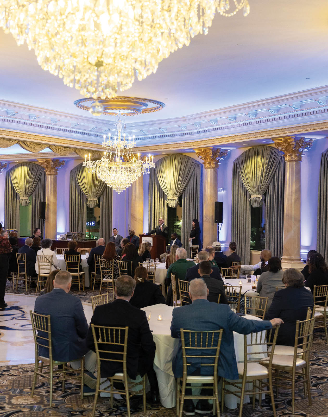 A group of people are sitting at tables in a large room with a chandelier hanging from the ceiling. The tables are set with wine glasses, forks, and knives. The people are engaged in conversation and enjoying their time together. AI generated content