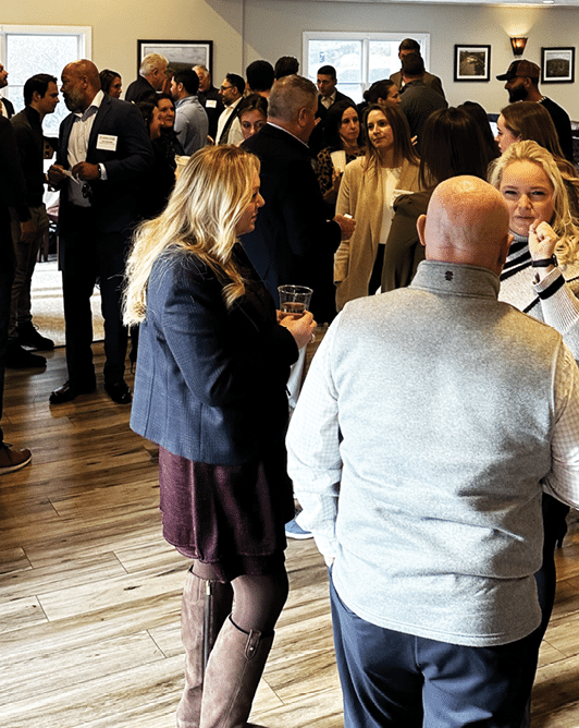 A group of people are standing in a room, socializing and enjoying drinks. There are several wine glasses and a cup visible in the scene. The people are engaged in conversation, creating a lively atmosphere. AI generated content