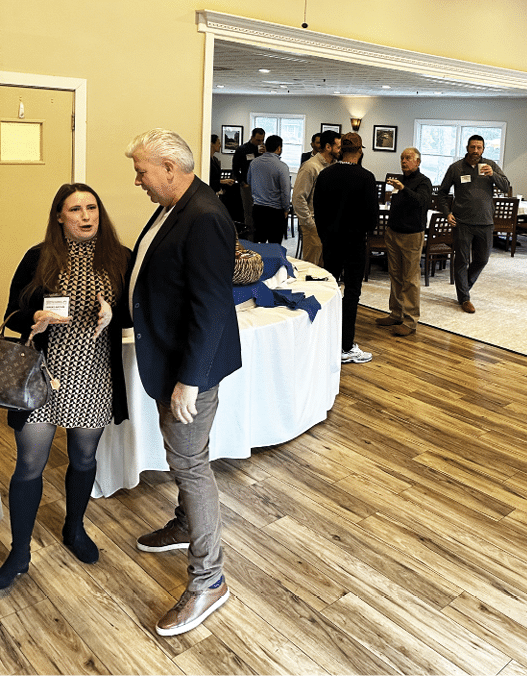 A group of people are gathered in a room, engaged in conversation. There are several chairs and a dining table in the room. A man and a woman are standing close to each other, talking. AI generated content