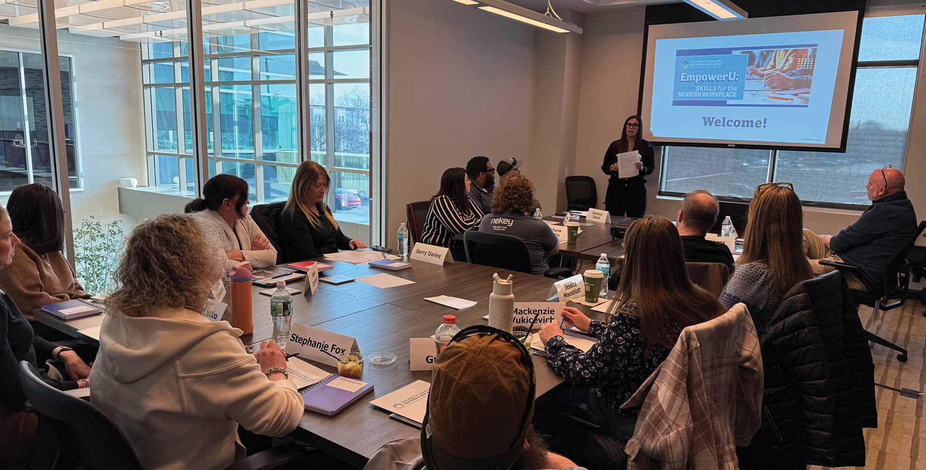 A group of people are sitting around a table in a conference room. A woman is standing at the front of the room, holding a paper. The attendees are engaged in a presentation. AI generated content