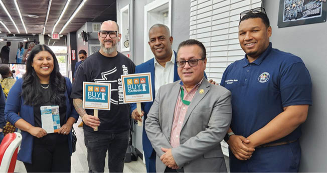 Five adults stand indoors holding signs and pamphlets promoting a buy local campaign in a well-lit room with modern decor. Description generated by AI
