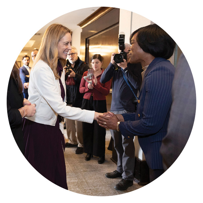 Two adults are shaking hands in a hallway while several people in the background take photos and observe. Description generated by AI