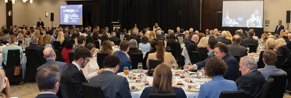 A large group of people seated at round tables in a banquet hall are attentively listening to a speaker at the front near projection screens. Description generated by AI