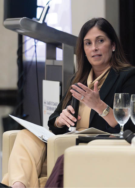 Two people are seated and engaged in a conversation, one wearing a black blazer and the other in a white jacket, with glasses of water on a table between them. Description generated by AI
