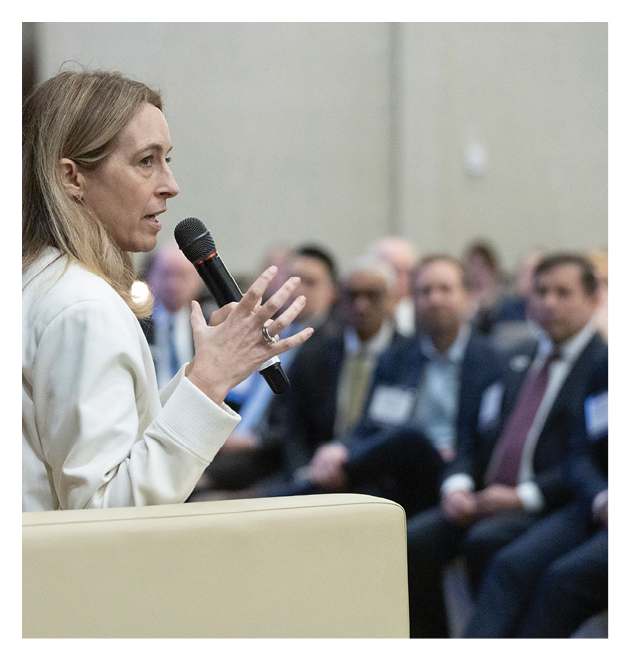 A person with long hair wearing a light-colored jacket is speaking into a microphone while seated on a beige chair in front of an audience. Description generated by AI