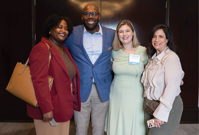 Four adults dressed in business casual attire stand closely together indoors, smiling and posing for a photo. Description generated by AI