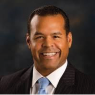 A person wearing a dark pinstripe suit, white shirt, and light blue tie is posed against a dark background. Description generated by AI