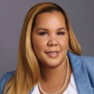 A person with long straight hair wearing a blue blazer, white blouse, and a pearl necklace is seated with hands clasped on a table against a plain gray background. Description generated by AI