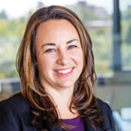 A person with long hair wearing a dark blazer and purple top is smiling in a bright office setting with glass walls and blurred background. Description generated by AI
