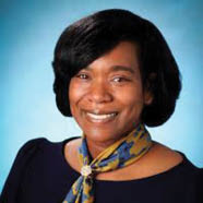 Person with short dark hair wearing a dark top and a yellow and blue patterned scarf against a light blue background. Description generated by AI