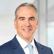 A person wearing a blue suit, white shirt, and light blue patterned tie is standing against a blurred neutral background. Description generated by AI