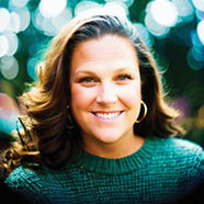 Person with long wavy hair wearing a green sweater and hoop earrings is standing in front of a background filled with circular bokeh lights. Description generated by AI