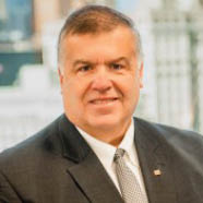 A person wearing a dark suit, white shirt, and patterned tie is standing in front of a blurred cityscape background. Description generated by AI