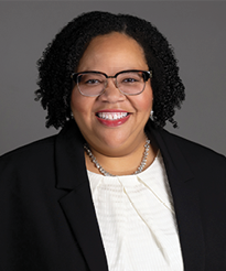 A person with curly hair wearing a black blazer over a white top and a silver necklace is posed against a plain gray background. Description generated by AI
